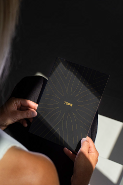 A person holds a black notebook with a gold sunburst design and the word &ldquo;TOPA&rdquo; in the center, outdoors on a sunny day.