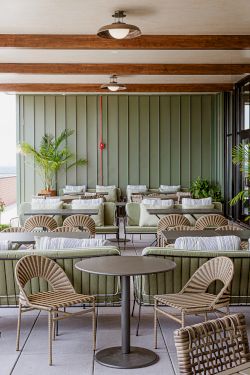 A stylish outdoor patio lounge with wicker chairs and small tables, green cushions, potted plants, and ceiling pendant lights.