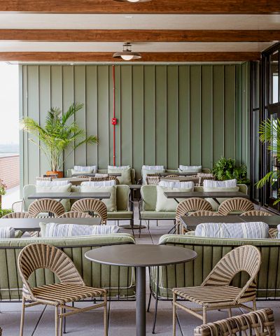 A stylish outdoor patio lounge with wicker chairs and small tables, green cushions, potted plants, and ceiling pendant lights.