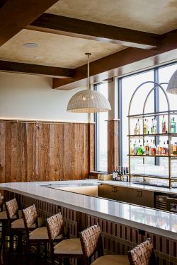 Modern bar interior with wood walls, a woven pendant light, liquor shelves, and chairs along the countertop.