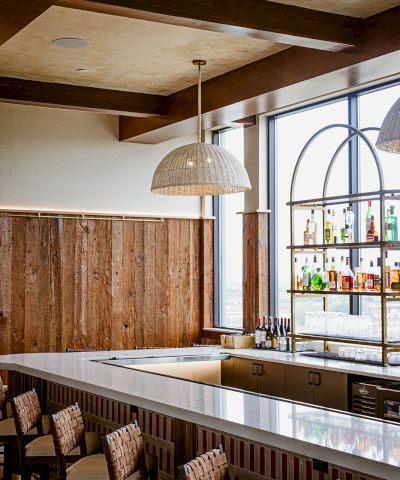 Modern bar interior with wood walls, a woven pendant light, liquor shelves, and chairs along the countertop.