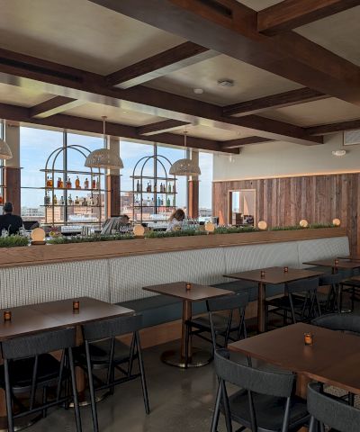 A stylish bar and dining area with wooden beams, a long counter with stools, and tables set for guests, backdrop of bottles and windows, cozy lighting.