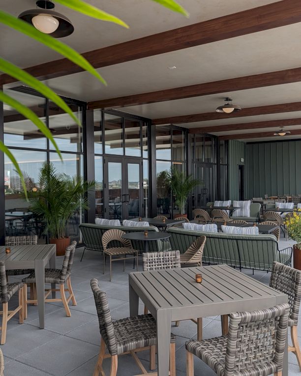 A modern outdoor patio with woven chairs and wooden tables under a pergola, potted plants, and comfortable seating along a shaded walkway.