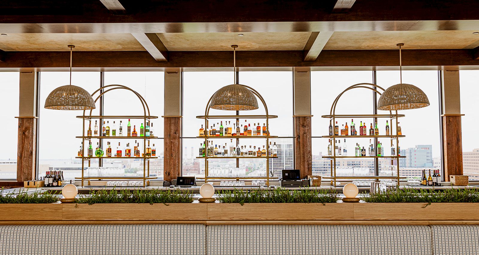 A rooftop-style bar with hanging woven lights and rows of colorful bottles behind a wooden counter by large windows.