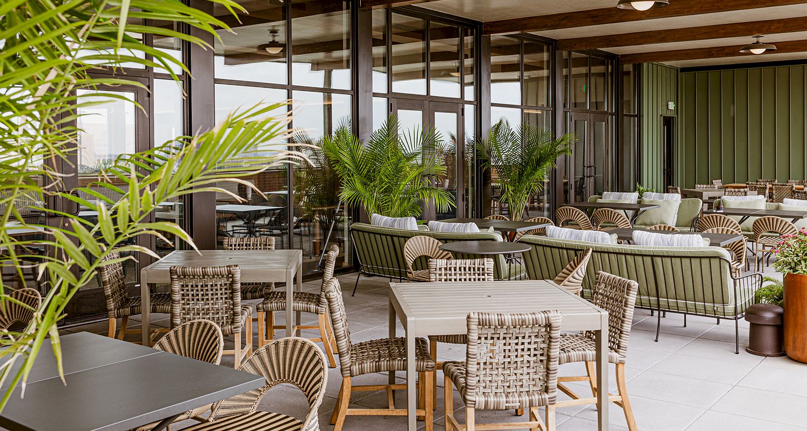 A modern outdoor patio with woven chairs and tables, tropical palms, and a glass corridor with wood beams.
