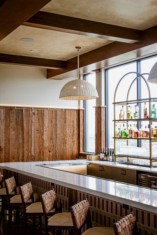 A modern bar interior with rustic wood walls, a pendant light, a drinks shelf of bottles, and high tables with chairs.