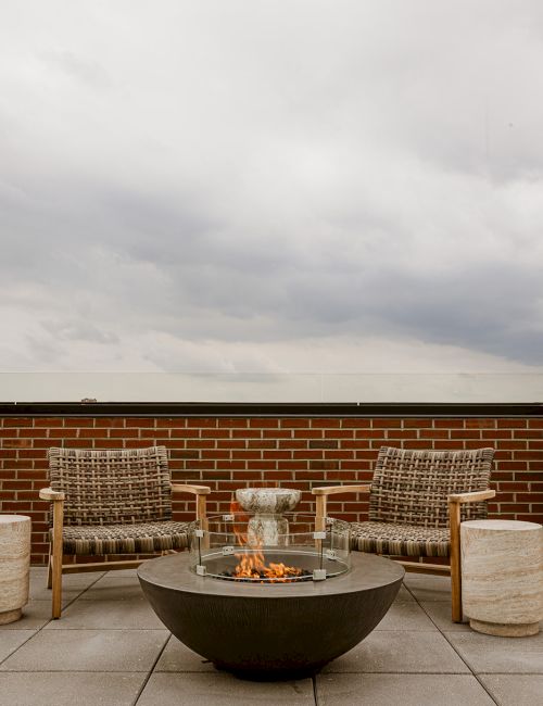 An outdoor patio with a glass fire pit burning, wicker chairs, stone stools, a brick wall, and cloudy sky above.
