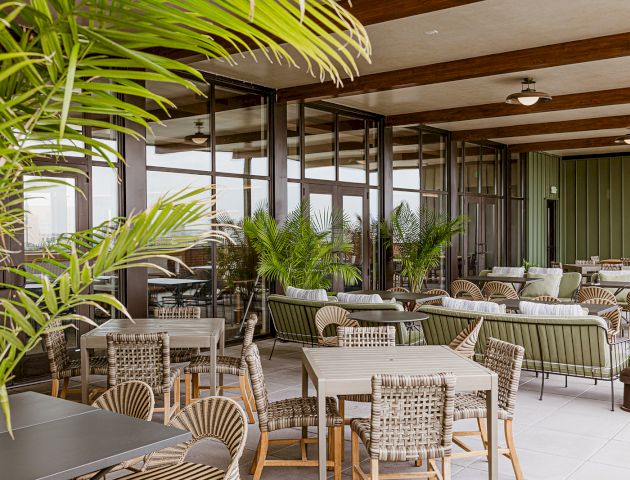 An indoor-outdoor patio with wicker chairs, tables, and lush potted palms behind glass walls and wood beams.