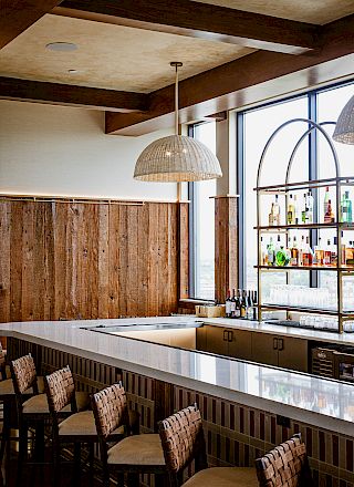 A modern bar interior with rustic wood walls, a long counter, bottles on shelves, and wicker chairs beneath a pendant lamp.