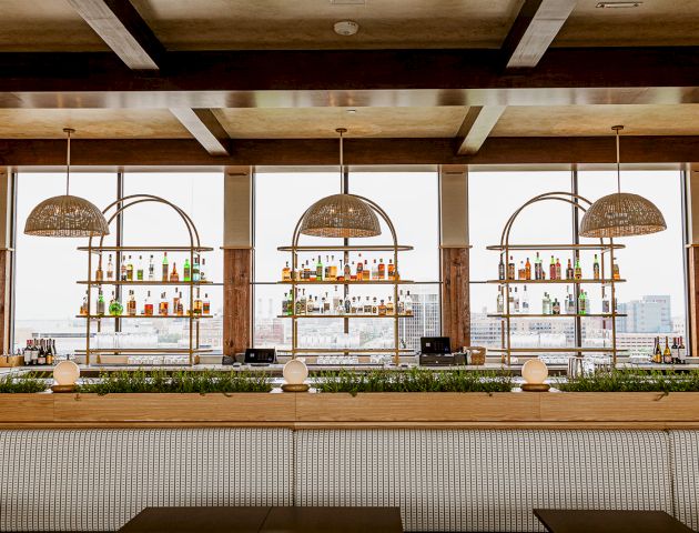 A modern bar lounge with shelves of colorful liquor bottles, hanging woven lamps, and a city skyline view through large windows.