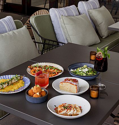 Outdoor patio scene with plush cushions and a table set with salads, appetizers, drinks, and candles.