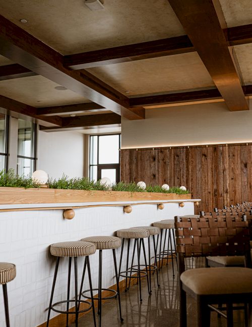 Modern bar interior with wooden beams, high stools, and a herb-lined counter under warm lighting.