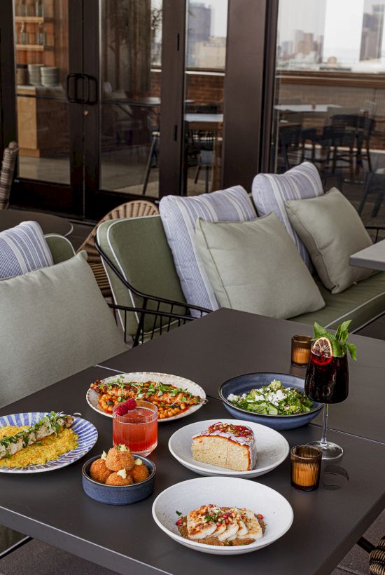 A sunny balcony patio with neatly arranged cushioned outdoor seating and a table full of plated appetizers, salad, drinks, and a dessert.