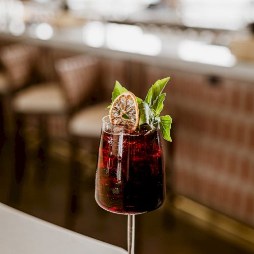 A dark red cocktail in a tall glass with ice, a citrus slice, and fresh mint leaves, served in a bar.