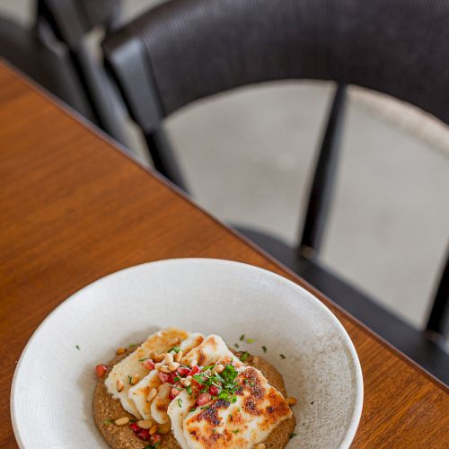 A bowl with savory flatbread topped with white cheese, herbs, pomegranate seeds, and pine nuts on a wooden table.
